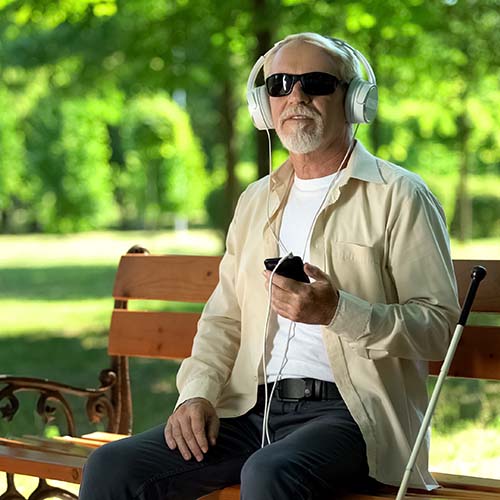 An apparent blind man in sunglasses sitting on a park bench wearing headphones and holding a phone. headphones.
