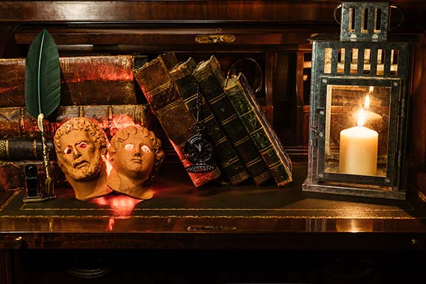 A spooky table with a pair of masks, some old books, and a lantern with a lit candle inside.